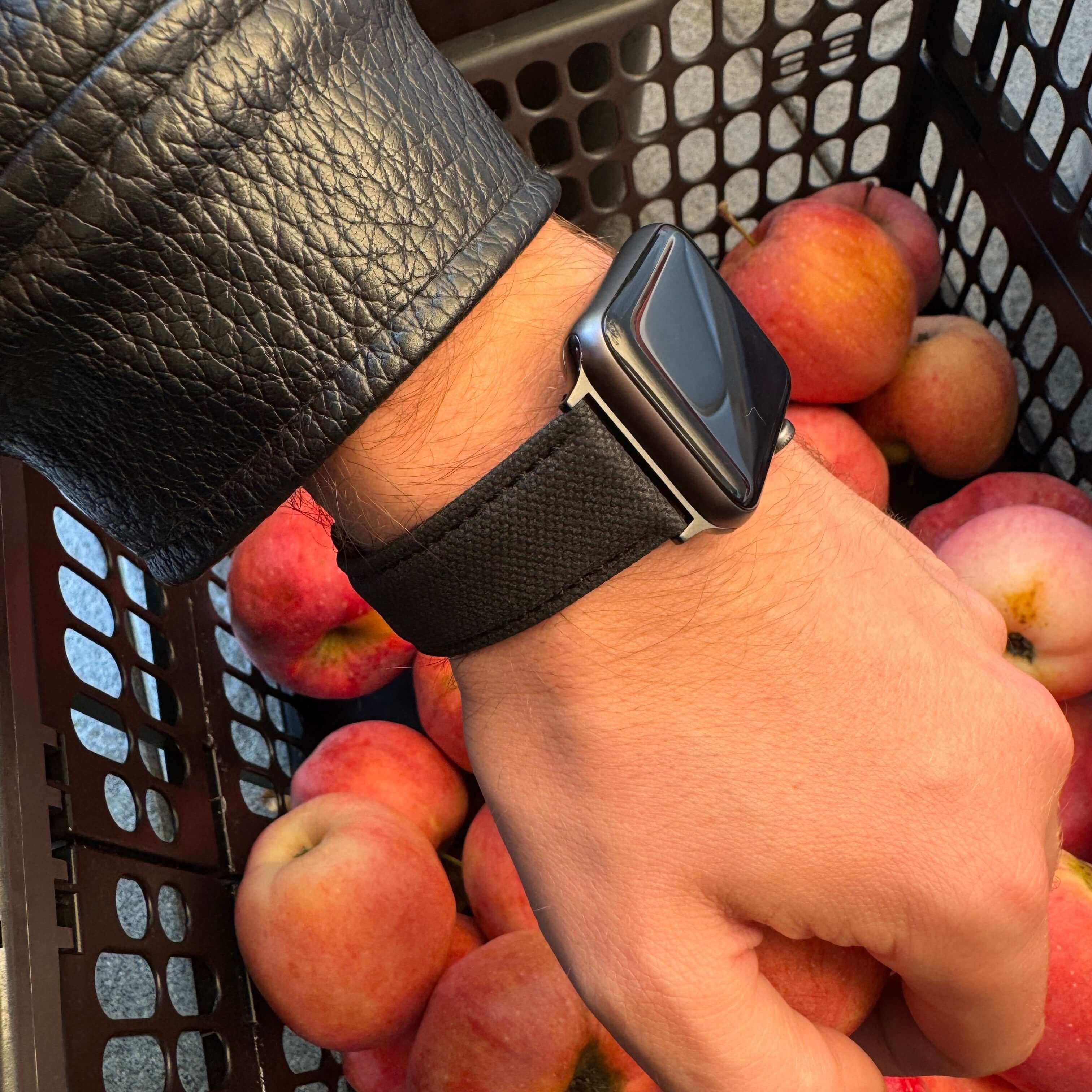 Schwarzes Canvas Apple Watch Band Baumwolle an einem Handgelenk, das einen Korb mit Äpfeln hält. Stilvoll und robust, ideal für Alltag und Business.