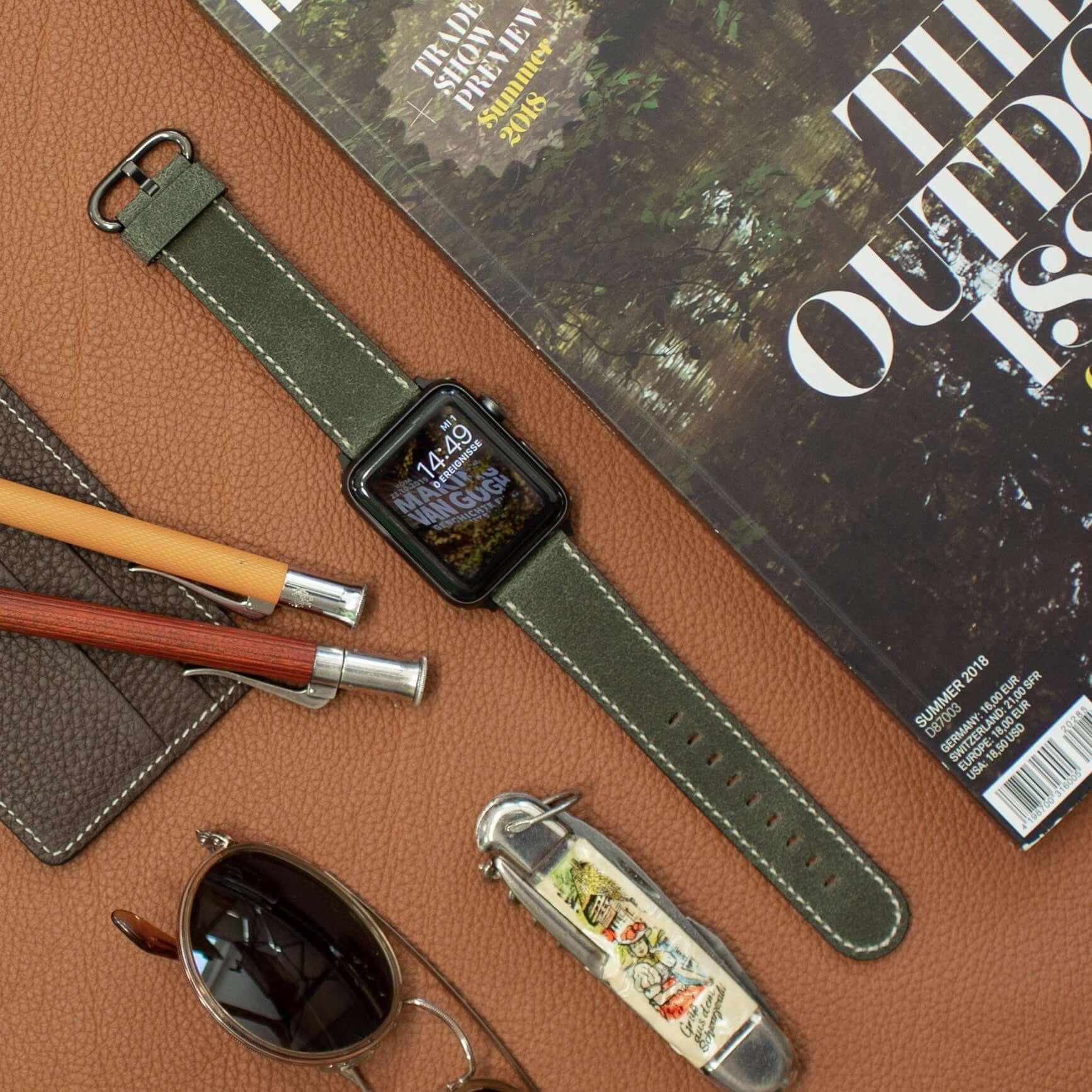 Apple Watch Lederarmband Vintage – Dunkelgrün auf Lederuntergrund mit Sonnenbrille, Messer und Zeitschrift, rustikales Ambiente.