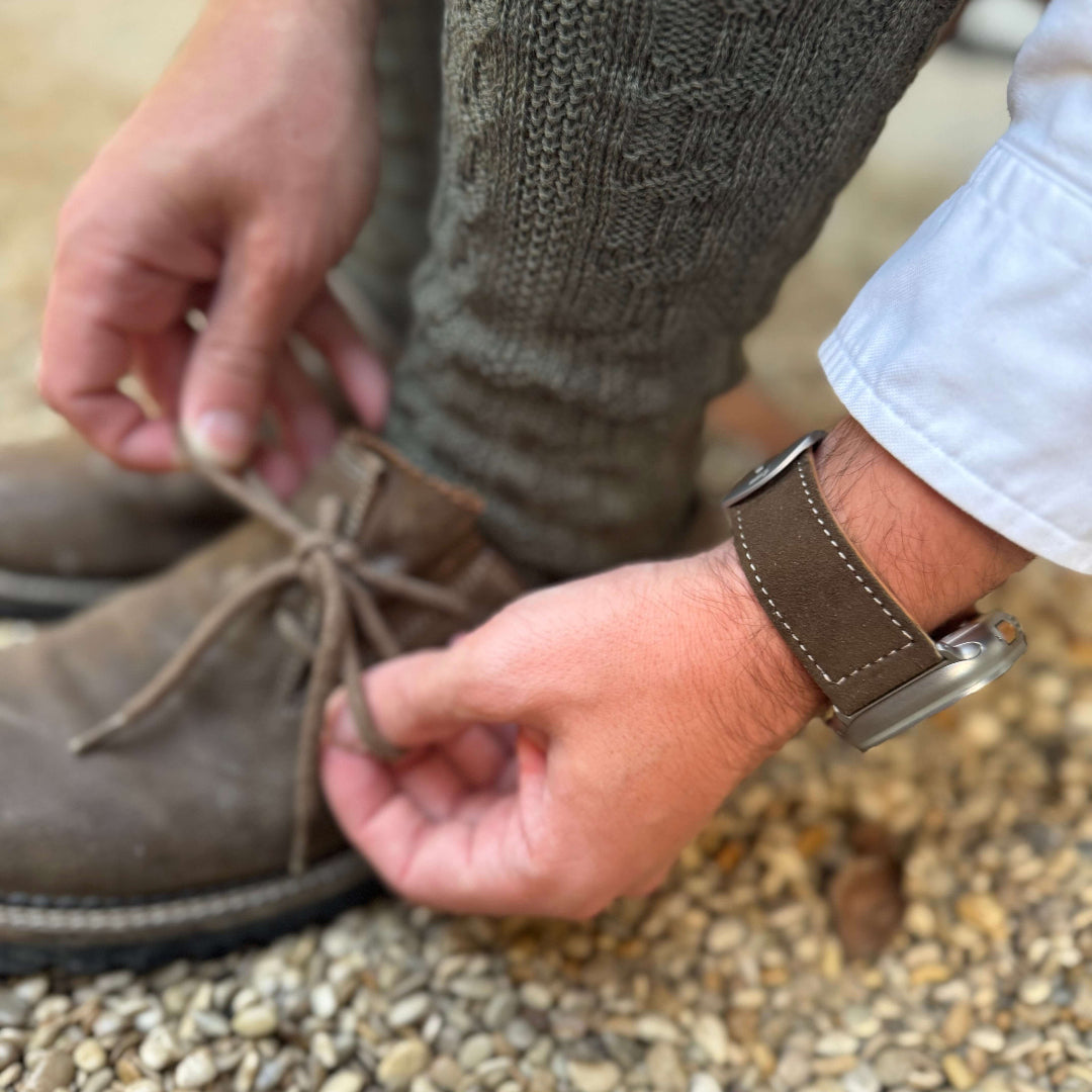 Hände binden Schuhe neben dunkelbraunes Apple Watch Ultra Lederhosen Armband – aus altsämisch gegerbtem Hirschleder, perfekte Wahl für Oktoberfest.