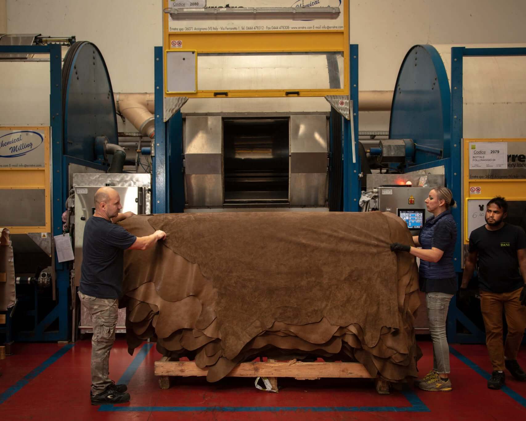 Workers handling leather sheets in a factory setting, with industrial machines in the background, showcasing the manufacturing process.