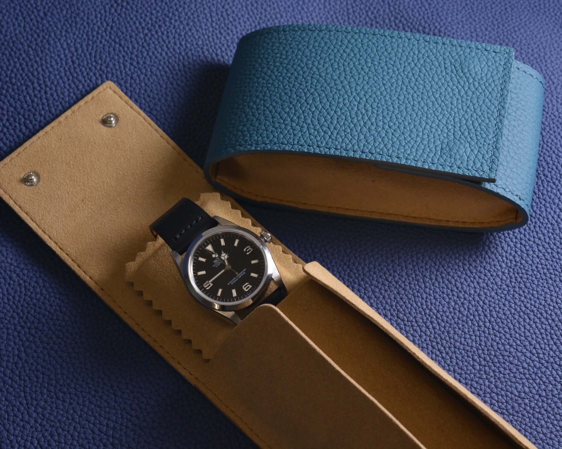 Luxury watch in a tan leather case next to a blue leather band on a textured background.