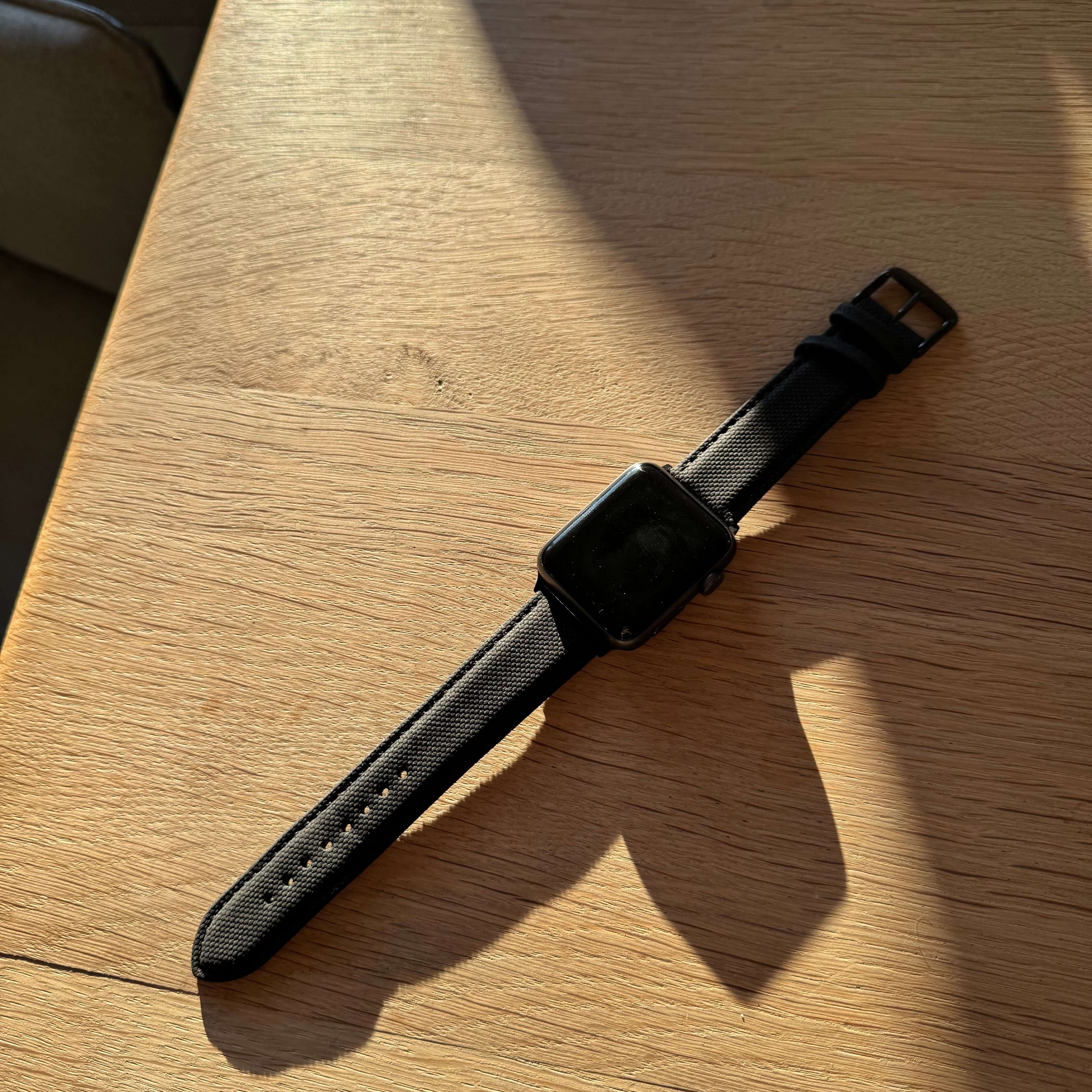 Schwarzes Canvas Apple Watch Band Baumwolle, stilvolles und robustes Uhrenband auf Holztisch mit Sonnenlicht.