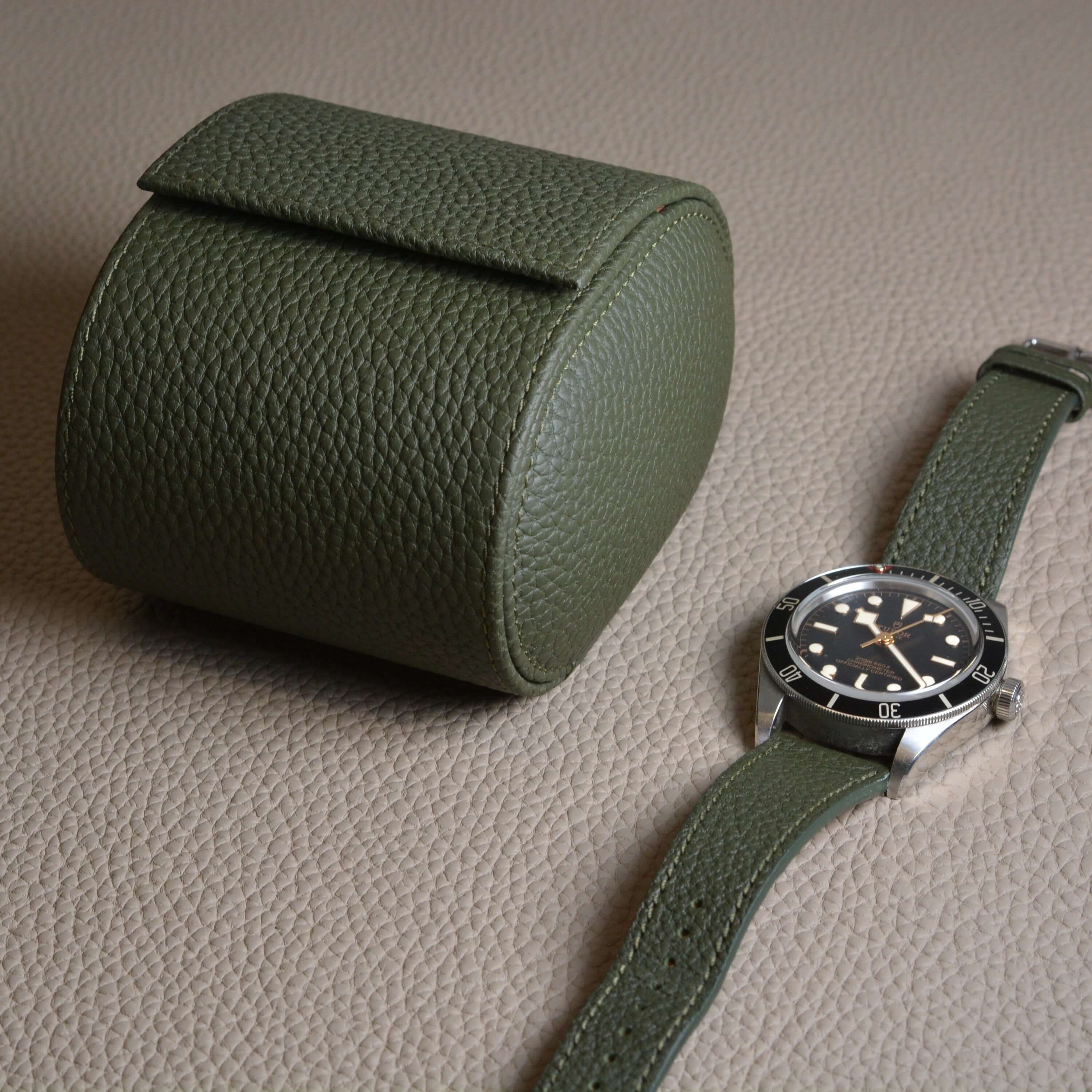 Olive leather watch roll and watch strap set on Togo leather background, featuring a classic black-faced watch.