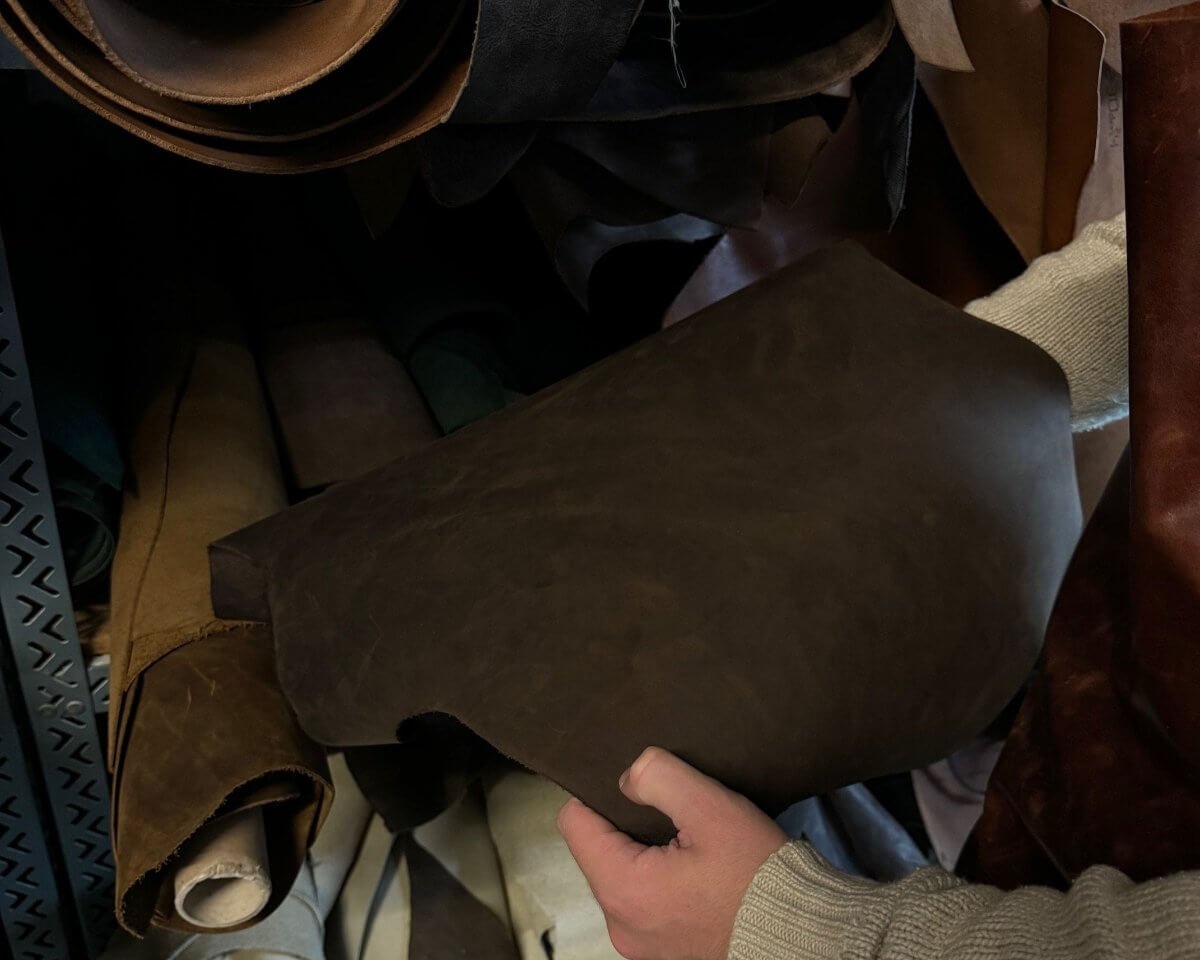 Nahaufnahme einer Person, die ein Stück hochwertiges dunkelbraunes Leder in einer Werkstatt hält, umgeben von verschiedenen zusammengerollten Lederhäuten in Braun-, Beige- und Schwarztönen. Perfekt für Lederhandwerk und Materialauswahl.