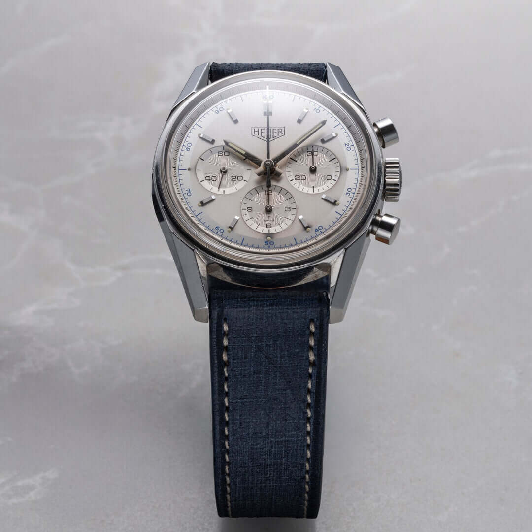 Luxuriöse Herrenuhr mit blauem Lederarmband, silbernem Zifferblatt und Chronographenfunktionsknöpfen, präsentiert auf heller Marmorfläche.