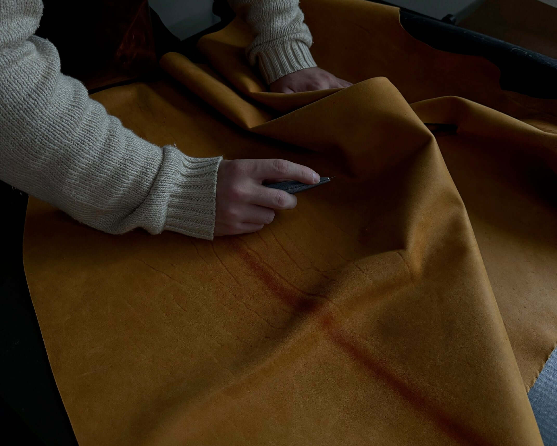 Person cutting leather fabric with a hand tool, wearing a beige sweater, focused on detailed work.