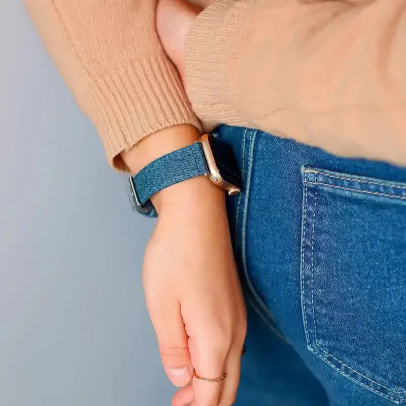 Close-up of person wearing a denim watch strap and brown sweater, resting hand on hip with blue jeans.