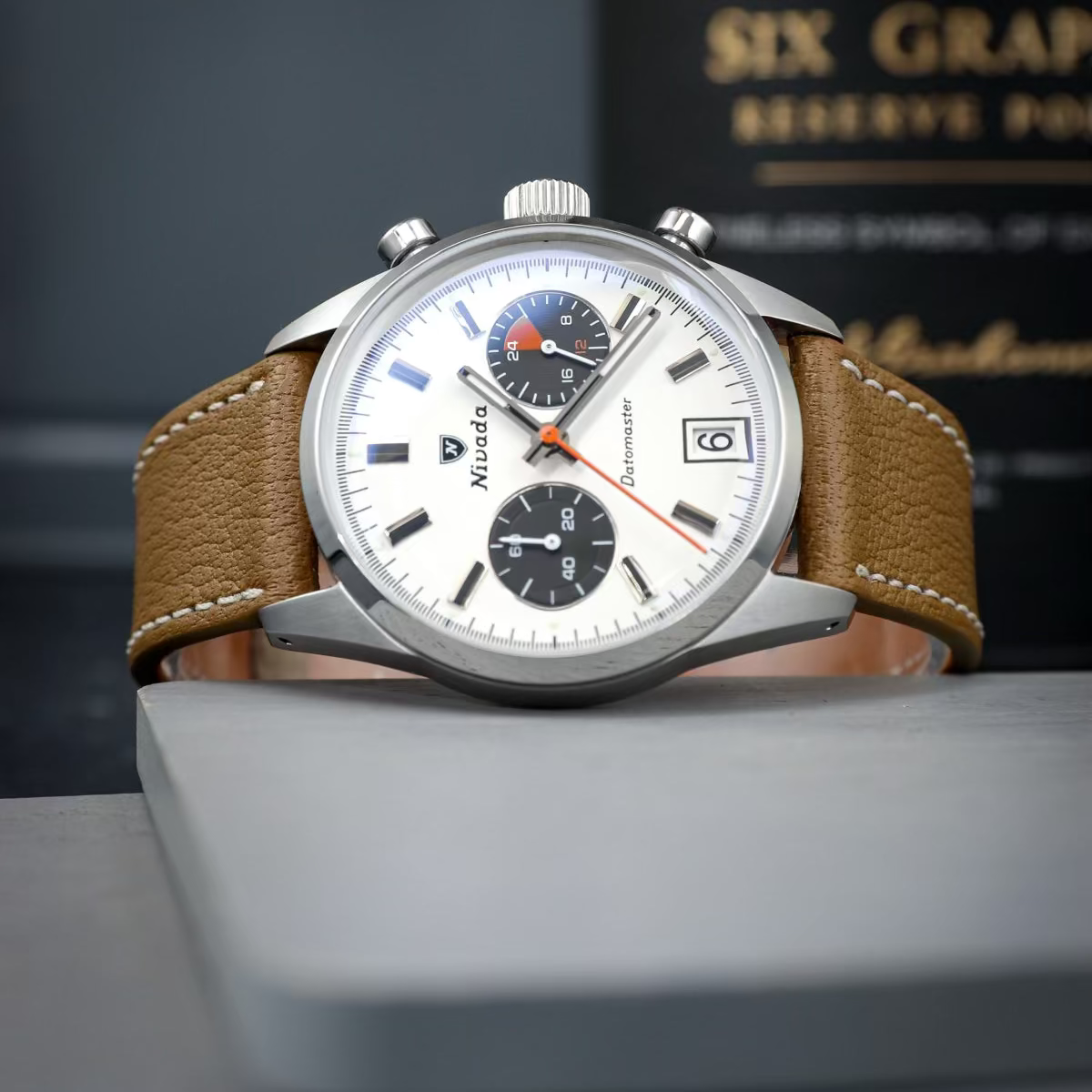 Elegante Uhr mit einem braunen, kurzen Leder Uhrenarmband. Die Uhr hat ein helles Zifferblatt mit schwarzen und roten Elementen, präsentiert auf einem grauen Untergrund.