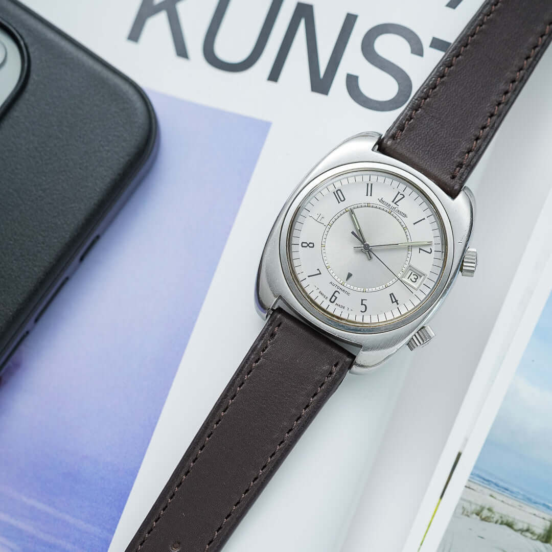 Jaeger-LeCoultre Memovox mit silbernem Zifferblatt und braunem Fluco Lederarmband, platziert auf einem Magazin mit moderner Typografie im Hintergrund.