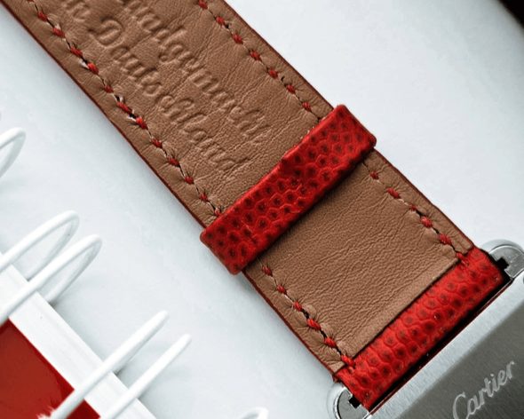 Nahaufnahme eines Armbands einer Cartier-Uhr. Das Armband besteht aus braunem Leder mit roten Nähten und roten Details aus strukturiertem Leder. Im Hintergrund ist ein rotes Notizbuch mit weißer Spiralbindung zu sehen. Auf dem Lederband ist eine Prägung auf Deutsch erkennbar.
