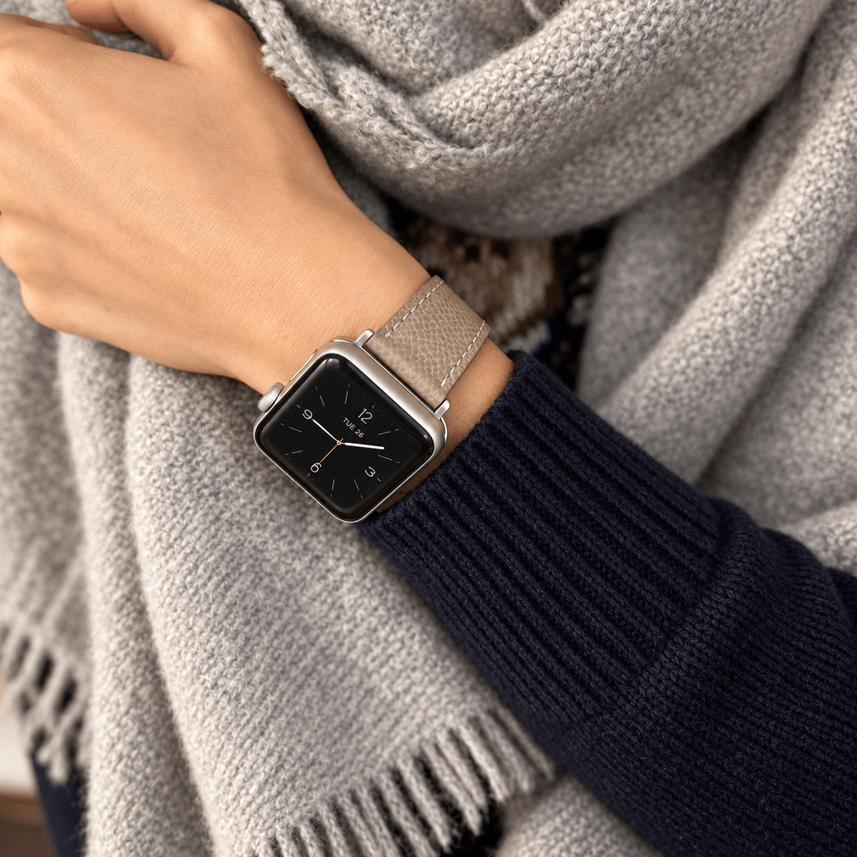 Luxuriöses Apple Watch Lederarmband Damen Cupertino Taupe an einer Hand, kombiniert mit einem grauen Schal.