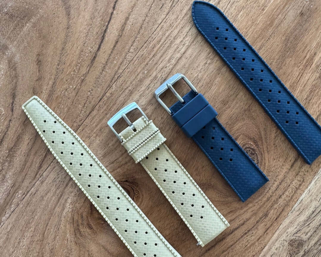 Vergleich von Silikon- und Kautschuk-Uhrenarmbändern auf einem Holztisch: zwei beigefarbene Silikonbänder mit feinem Muster und ein dunkelblaues Kautschukband mit Perforierung und glatter Oberfläche.