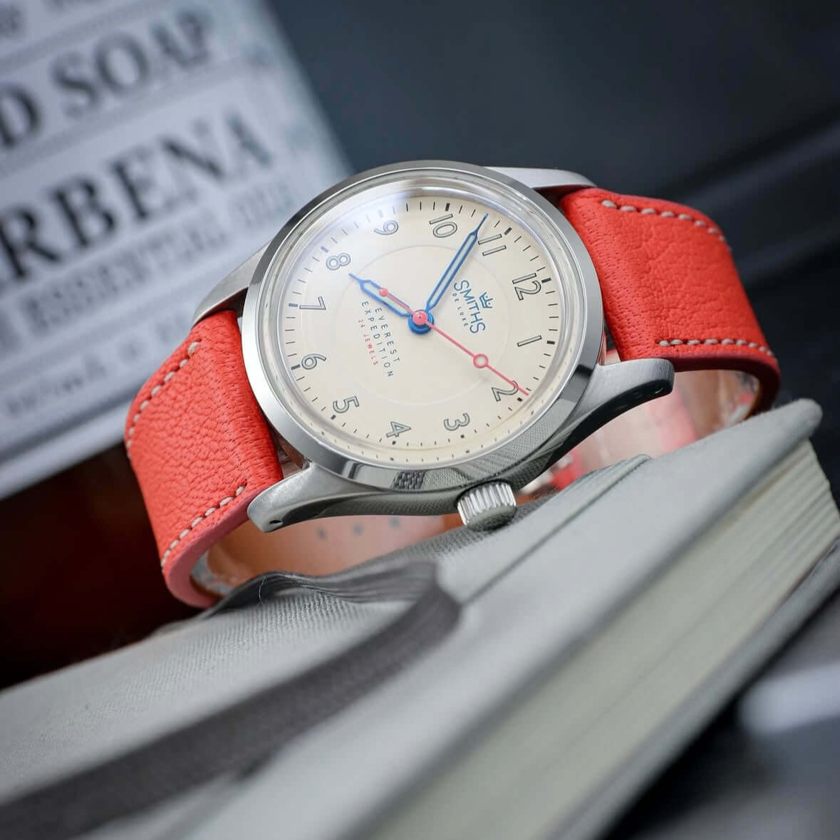 Biarritz Uhrenarmband in Korallenrot an einer stilvollen Uhr, die auf einem offenen Buch liegt, umgeben von einem modernen Ambiente.
