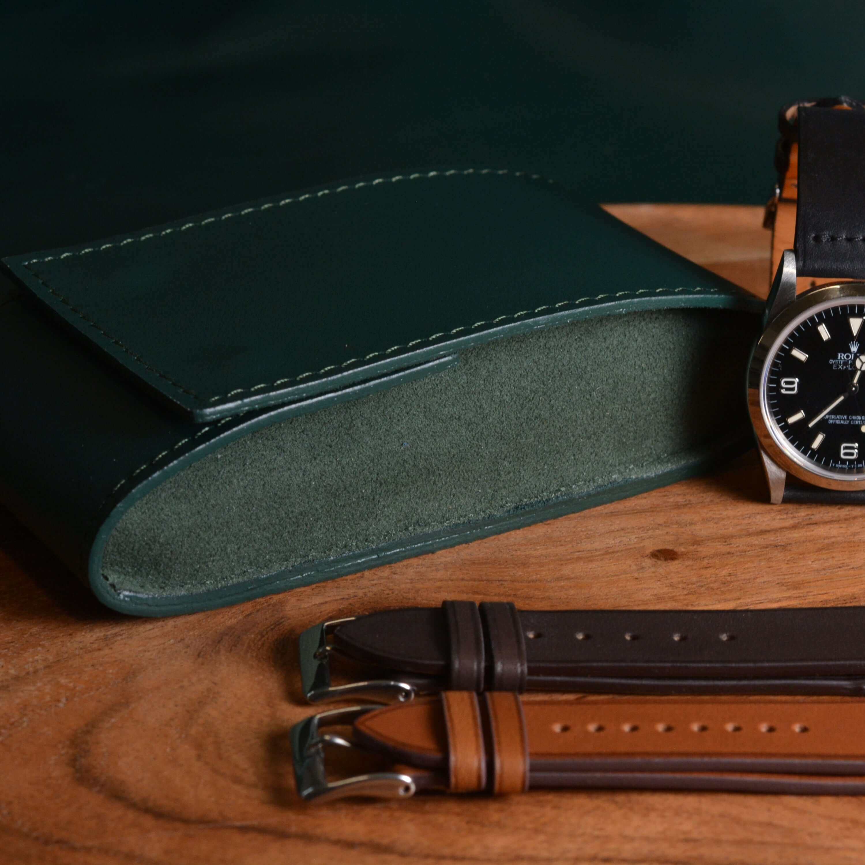 Hochwertige Uhrenaufbewahrung in Grün neben einer Rolex mit schwarzem Lederband – stilvolles Reiseetui aus italienischem Leder mit weichem Alcantara, maßgeschneidert für Premiumuhren.