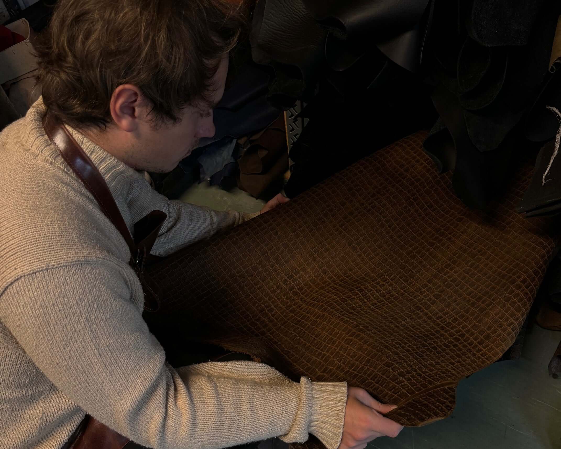 Person examining brown leather sheet in workshop, wearing a cream sweater and apron. Leather craft enthusiast in action.