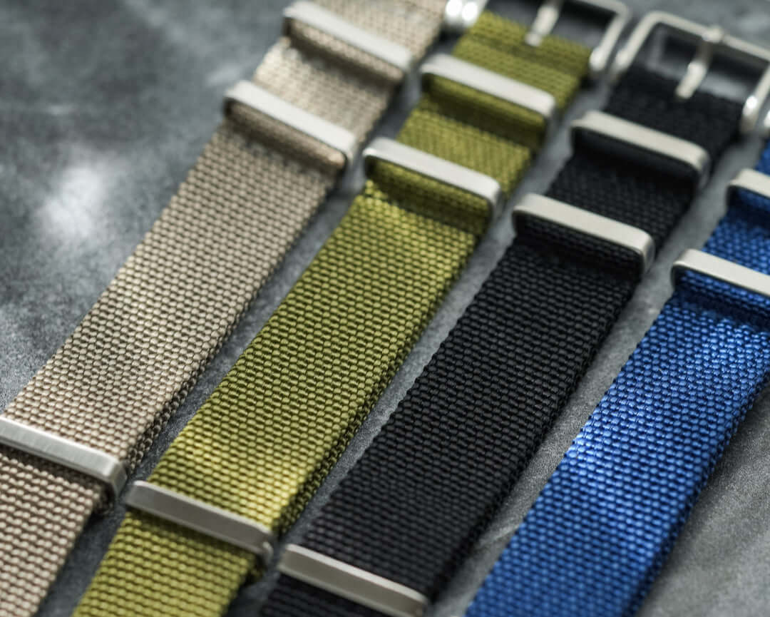 Vier verschiedene NATO Bänder nebeneinander – Detailaufnahme von vier strapazierfähigen NATO Bändern in unterschiedlichen Farben und Designs, einschließlich des grünen NATO Bands mit Fluco Logo