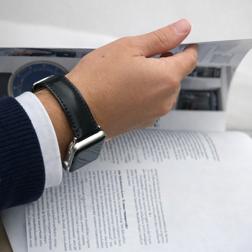 Apple Watch Armband Luxus aus Horween Shell Cordovan in Schwarz am Handgelenk, während eine Zeitschrift gelesen wird.