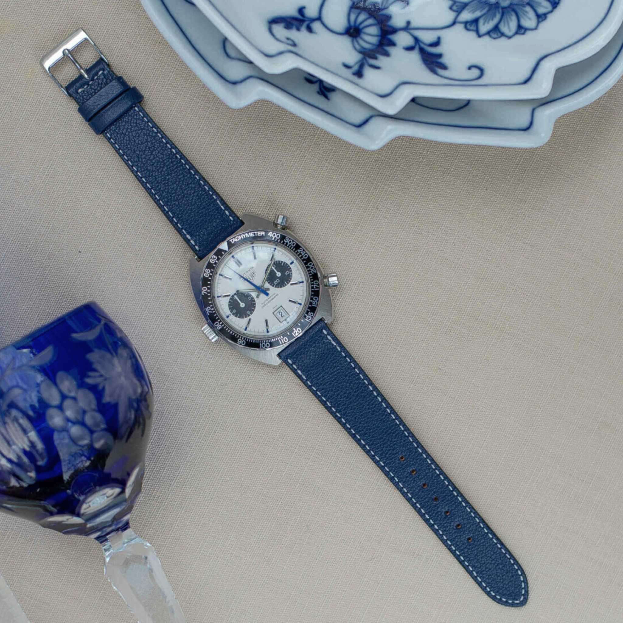 Flache Ansicht einer Uhr mit einem Blaues Ziegenleder Uhrenarmband, abgebildet neben einem eleganten blauen Glas und dekorativem Geschirr.