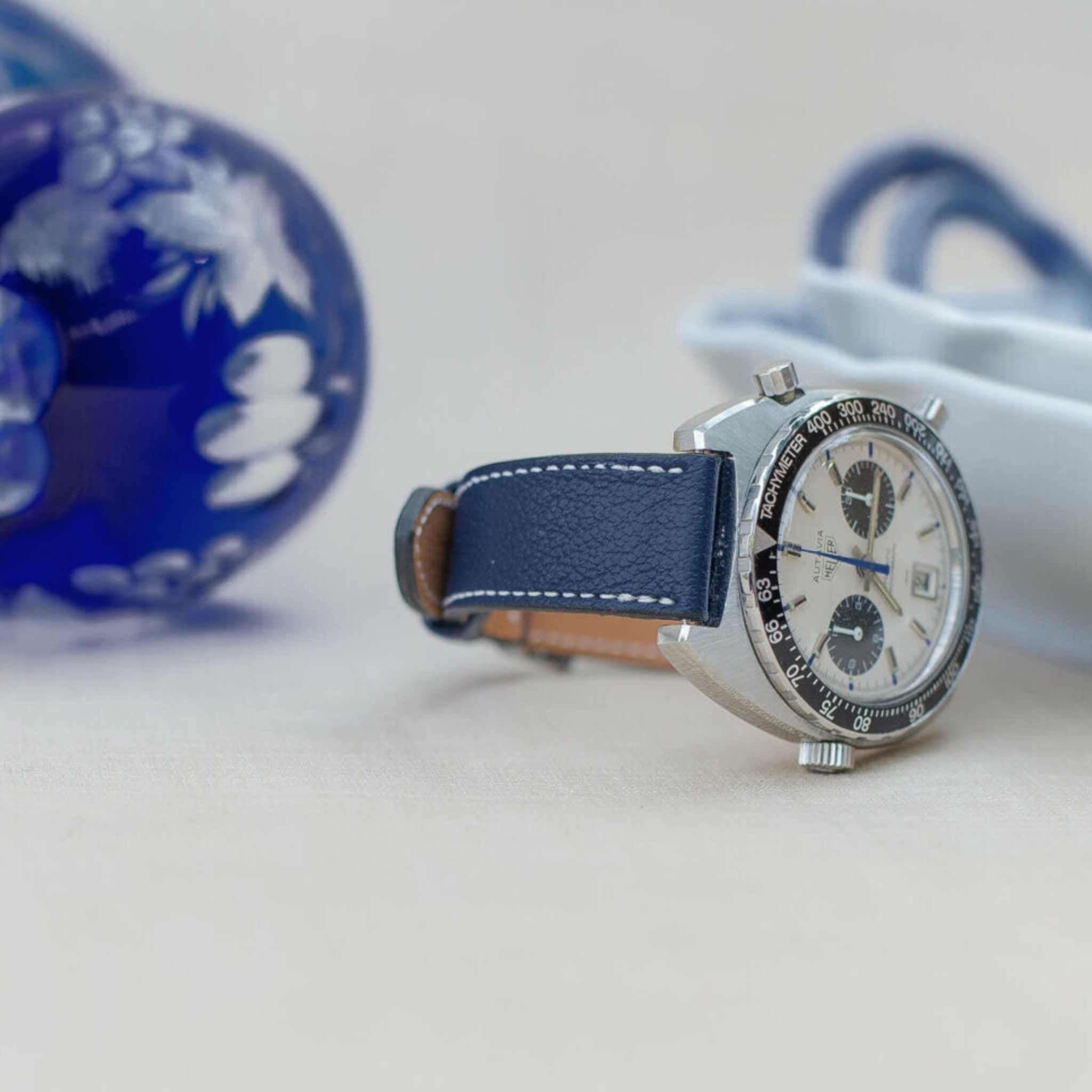 Nahaufnahme einer Uhr mit einem Blaues Ziegenleder Uhrenarmband, hellen Ziffern und Chronographen-Funktionen, im Vordergrund mit einem blauen Glasobjekt im Hintergrund.