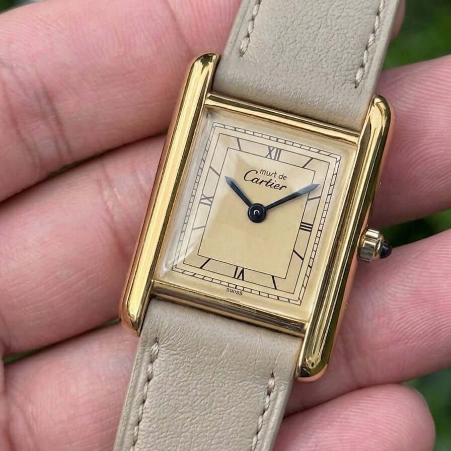 Hand holding a classic Cartier watch featuring a tan dial and beige leather strap.
