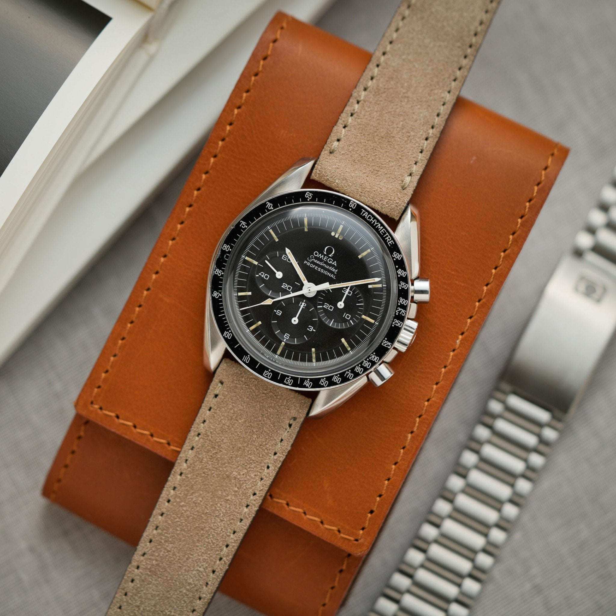 Beiges Nizza Velourslederarmband an einer Omega Speedmaster, von oben fotografiert und auf einem 1er Barenia Case liegend, elegantes Uhrenarmband aus hochwertigem Veloursleder.
