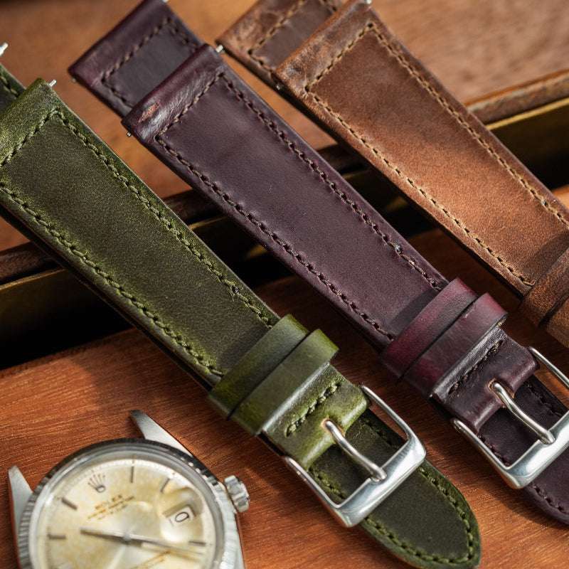 Bordeauxfarbenes, grünes und braunes Horween Chromexcel Uhrenarmband nebeneinander, im Hintergrund eine Rolex Uhr – Detailaufnahme von drei exklusiven Uhrenarmbändern aus hochwertigem Chromexcel Leder, perfekt für sportliche und luxuriöse Uhren