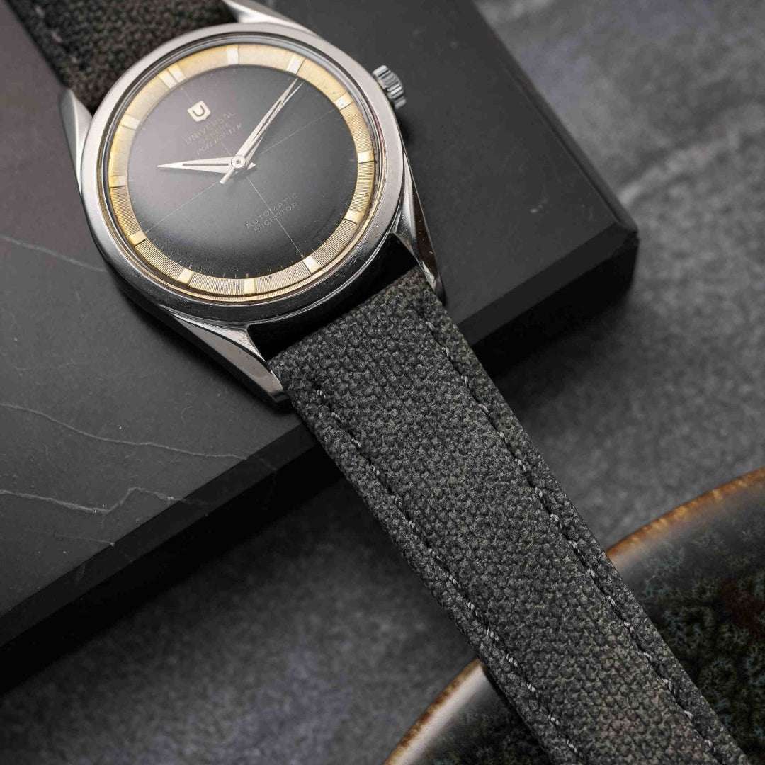 Universal Genève Polerouter mit grauem Canvas-Uhrenarmband, präsentiert auf einer schwarzen Marmorplatte, edler Vintage-Stil.