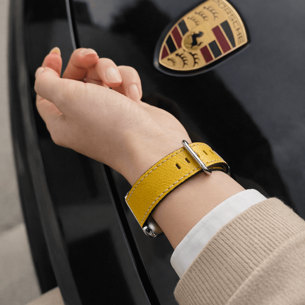 Luxuriöses Apple Watch Lederarmband Damen Cupertino Gelb am Handgelenk mit Porsche Logo im Hintergrund.