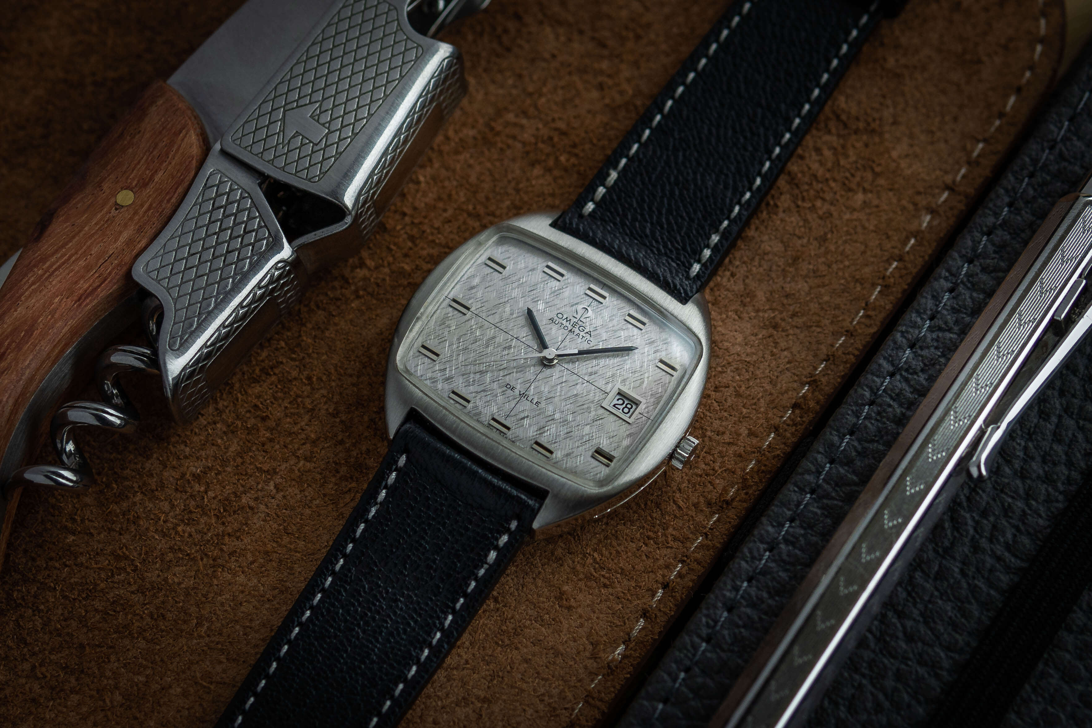 Schwarzes Lederarmband einer Omega-De-Ville-Vintage-Uhr mit sichtbarer feiner Naht, liegend auf braunem Untergrund.