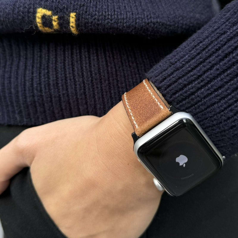 Ein Handgelenk mit einem braunen Vintage Apple Watch Lederarmband, das die Apple Watch 44mm ziert. Das modische Design passt perfekt zu lässiger Kleidung.