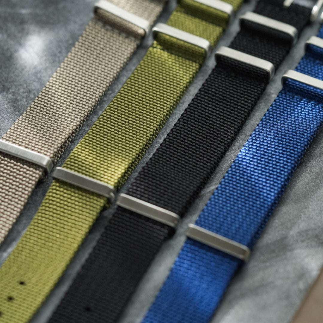 Vier verschiedene NATO Bänder nebeneinander – Detailaufnahme von vier strapazierfähigen NATO Bändern in unterschiedlichen Farben und Designs, einschließlich des grünen NATO Bands mit Fluco Logo